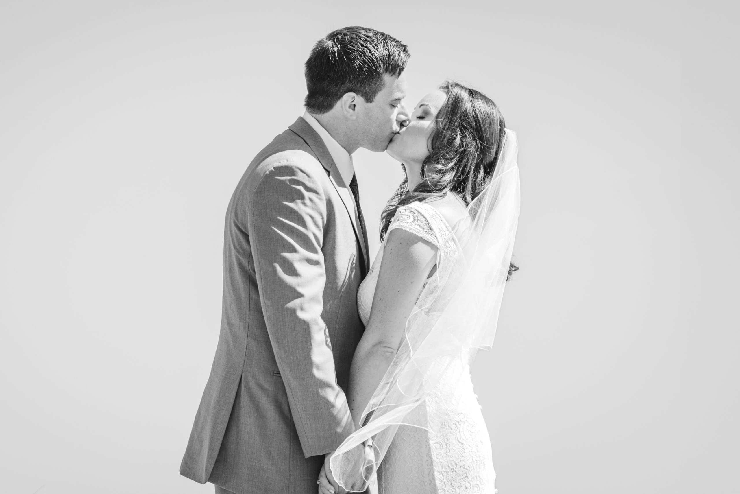 A black and white photo of a bride and groom sharing a sweet kiss after their wedding with a clear sky behind them.