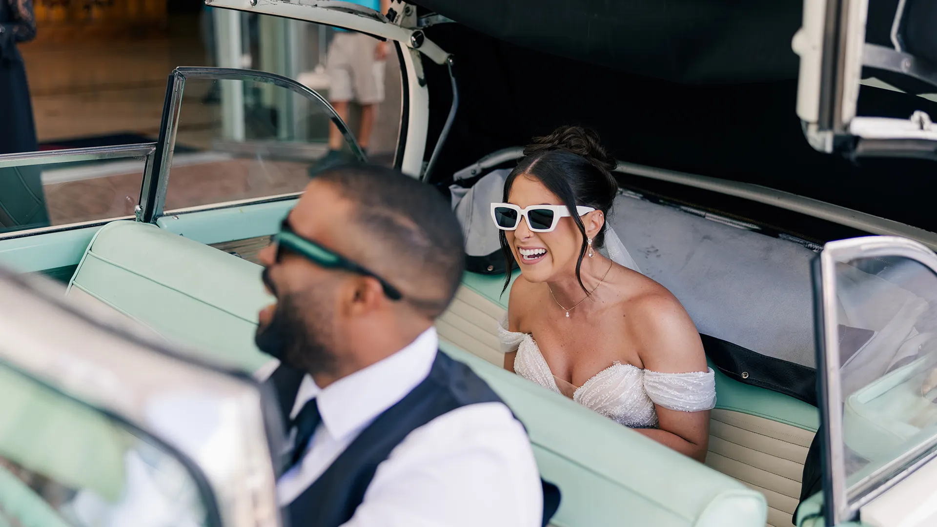 A bride sitting in the rear seat of a classic car laughing with the driver of the car.