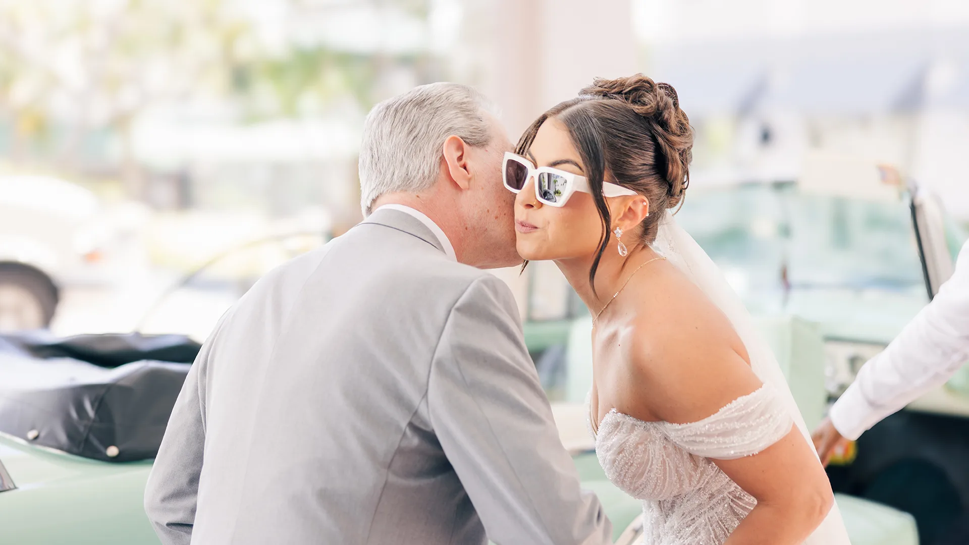 A bride being escorted by her father and sharing a kiss on the cheek before she enters the beautiful classic car and departs for her first look.