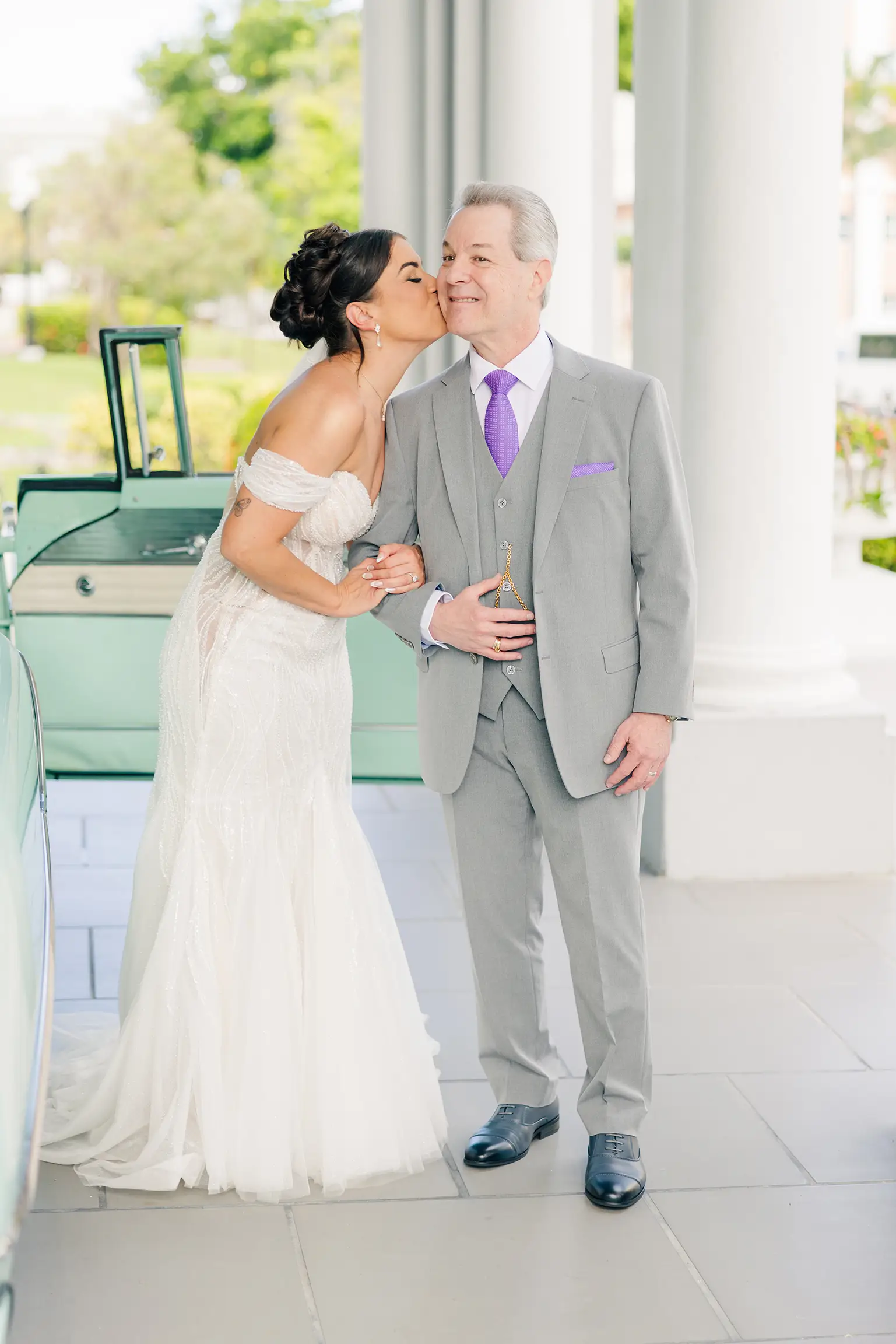 A bride giving her father a kiss on the cheek outside next to a classic car smiling and exited for her wedding day.