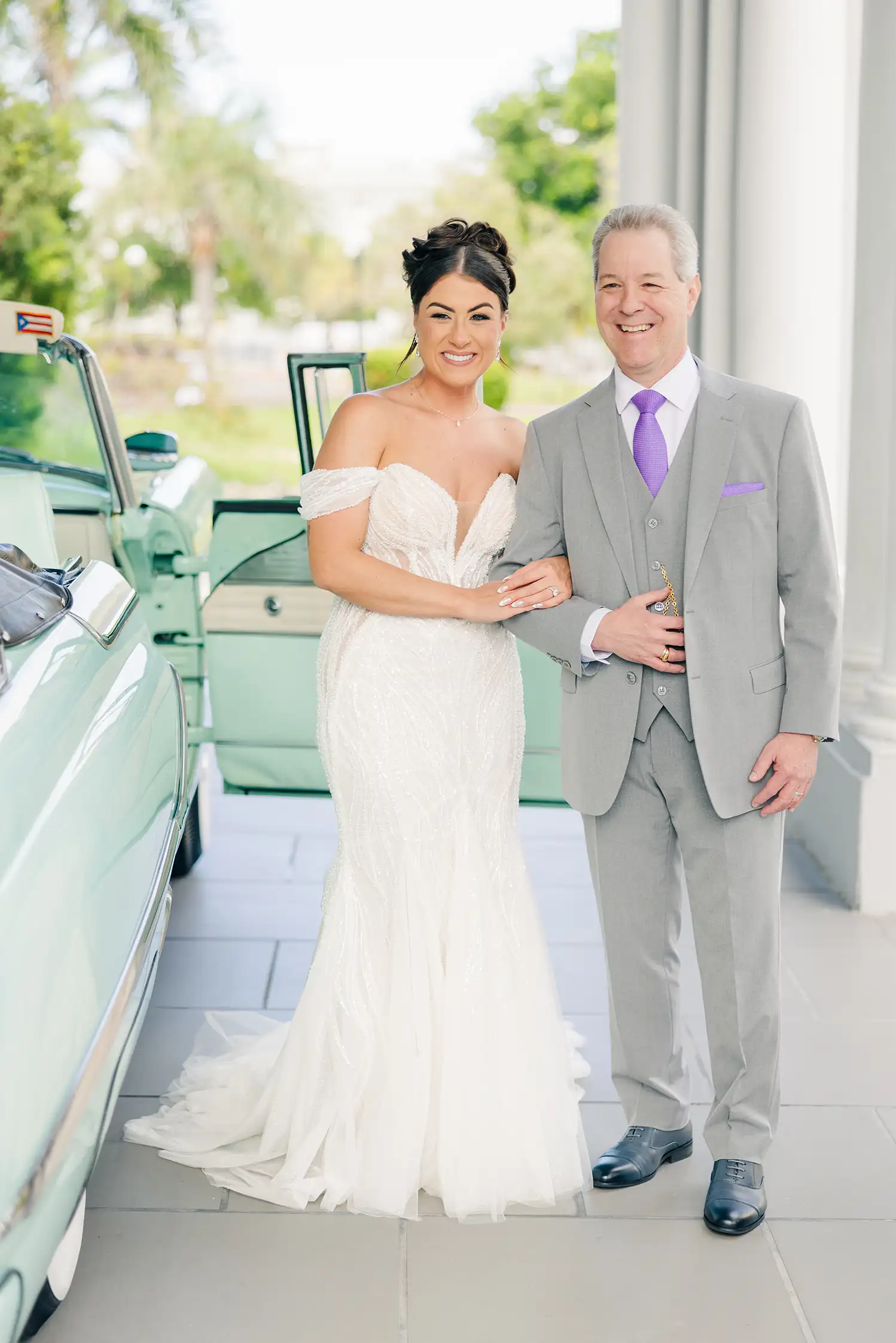 A bride leaning her head on her fathers shoulder outside next to a classic car smiling and exited for her wedding day.