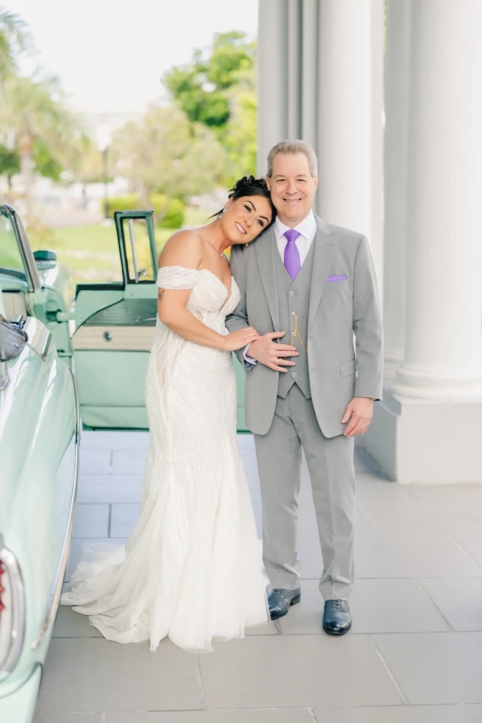 A bride with her father being escorted out of a classic car on her wedding day posing with her dad on her fathers shoulder smiling.