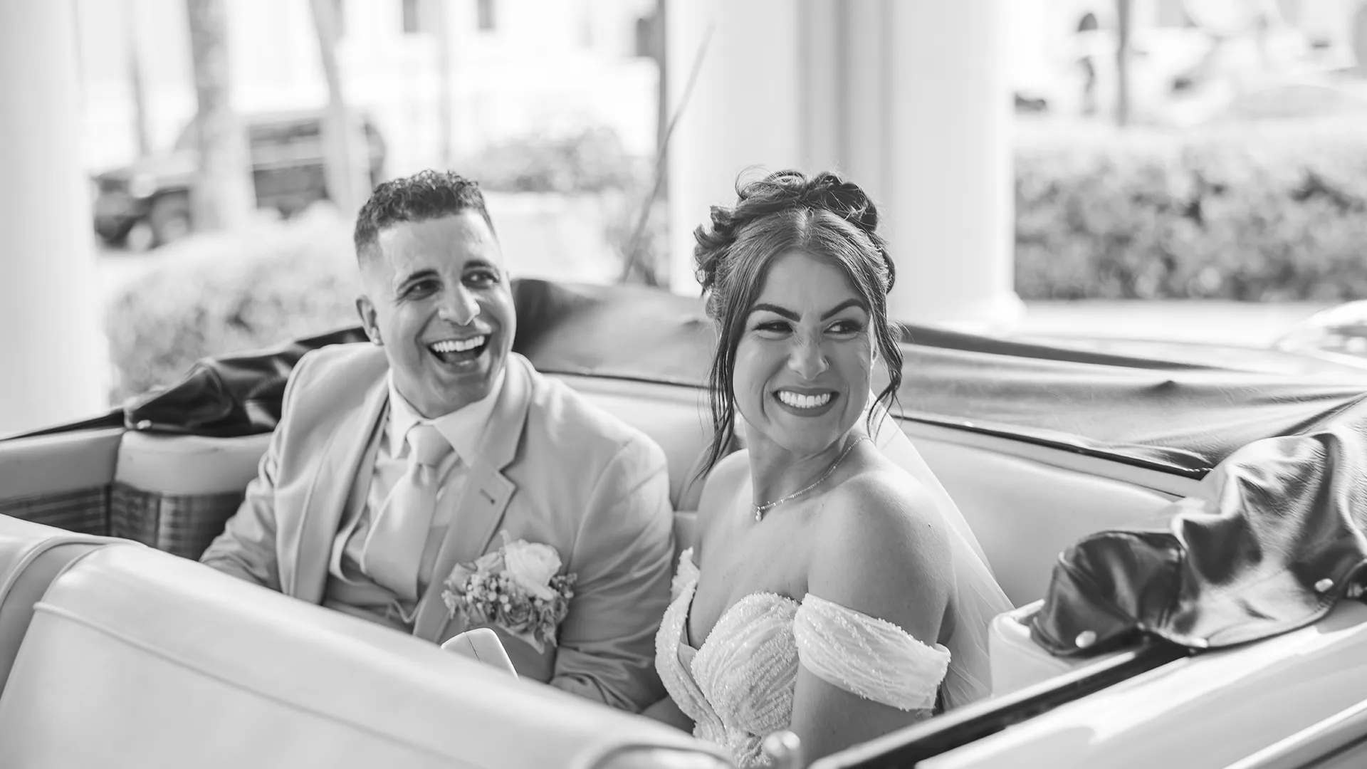 A bride and groom smiling at their guests in the back seat of a classic car arriving to their wedding ceremony after their first look.
