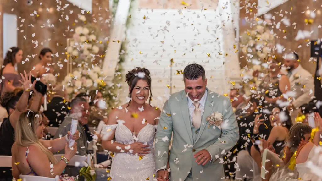 The bride, Laura and groom, Eddie (WWE referee) smiling while leaving their wedding ceremony with confetti flying in the air, guests cheering for them.