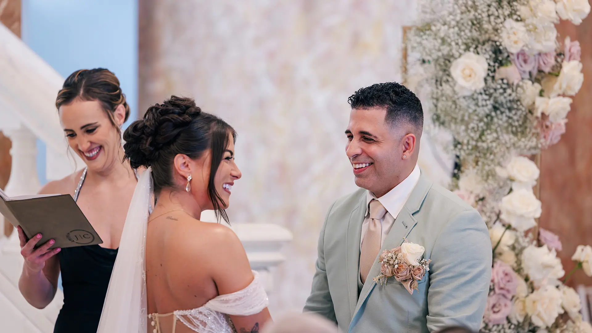 A bride and groom at the alter laughing at the officiant's speech with beautiful flower arches being them.
