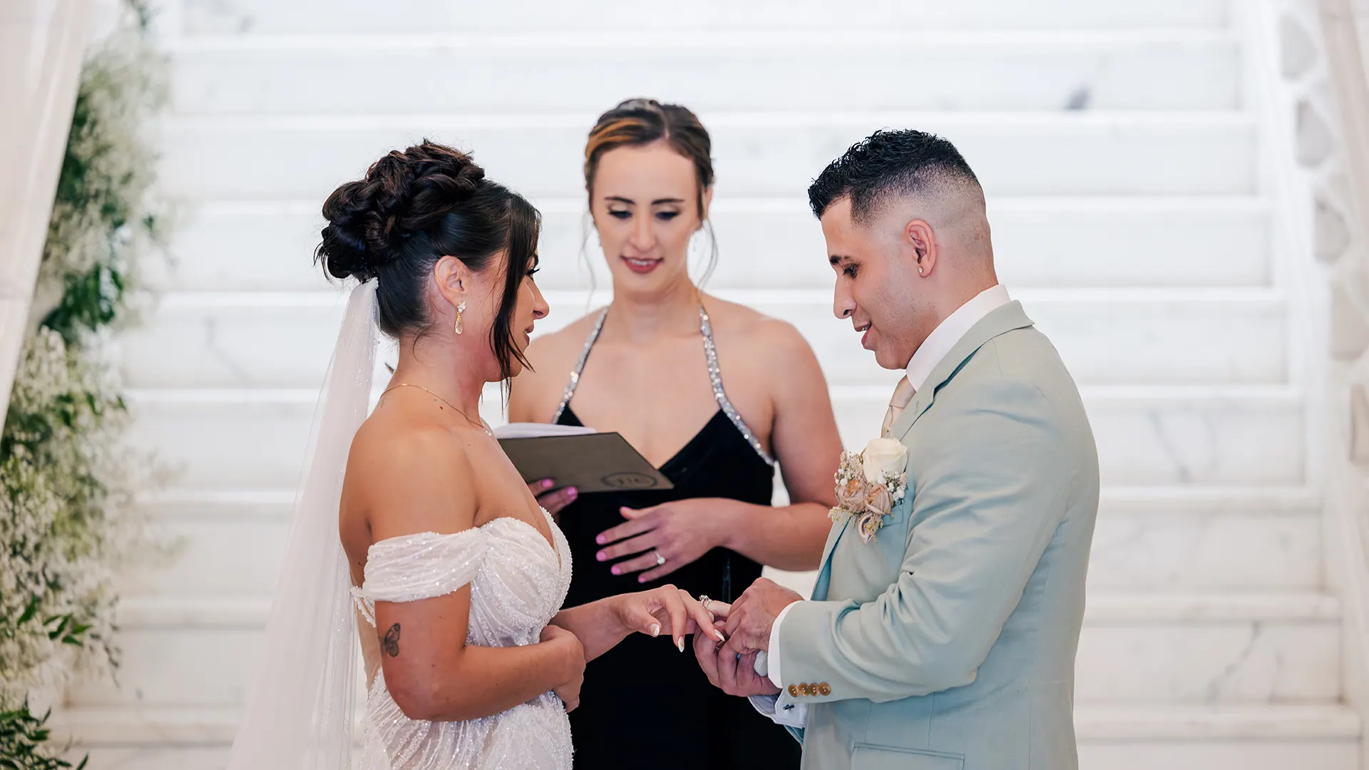 The groom placing the ring on the bride's finder with the officiant at a beautiful alter with white flowers and a white marble staircase.
