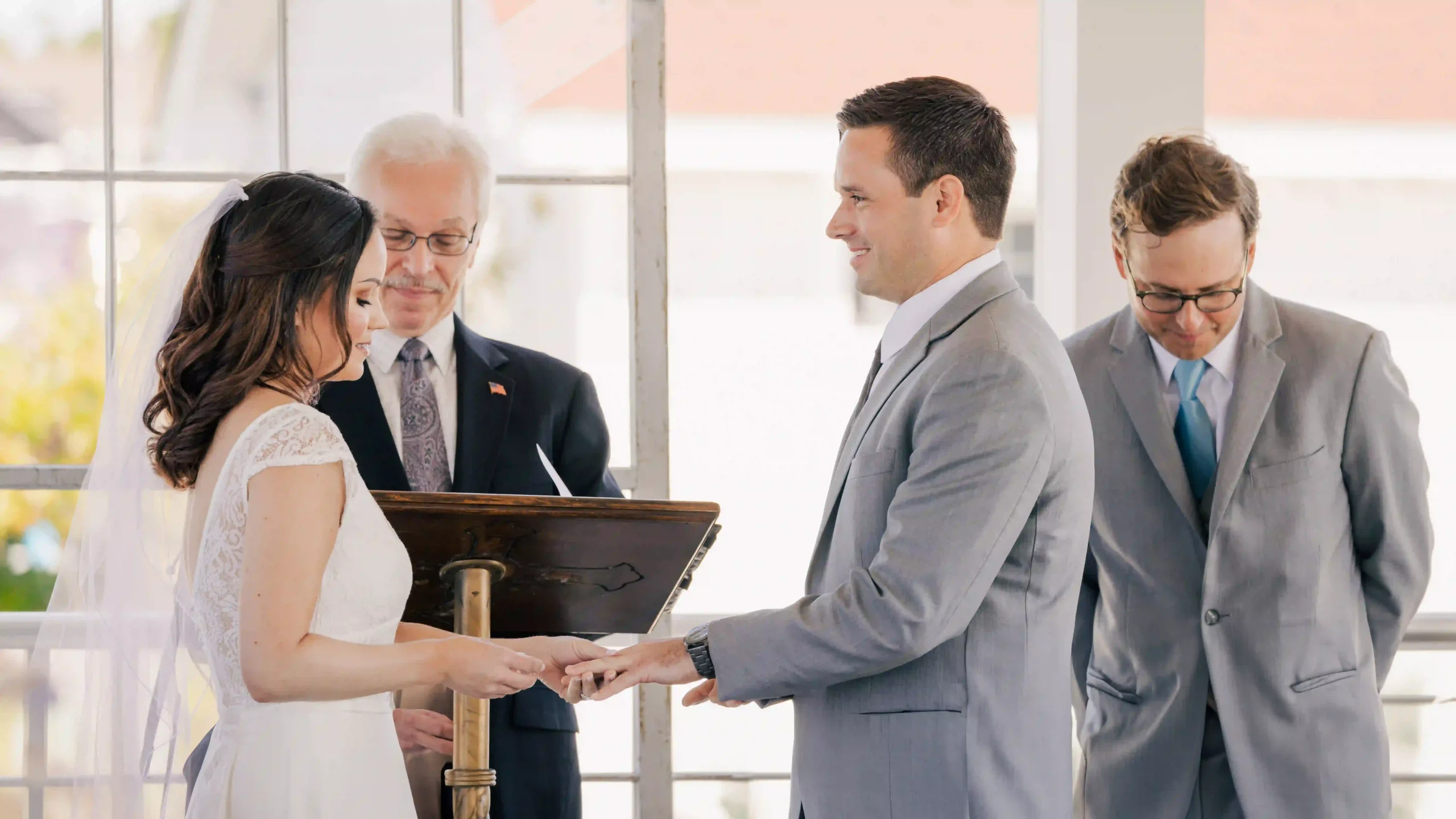 Bride smiles lovingly as she places a wedding ring on the groom's finger during their wedding ceremony at the altar with officiant and best man in the background.