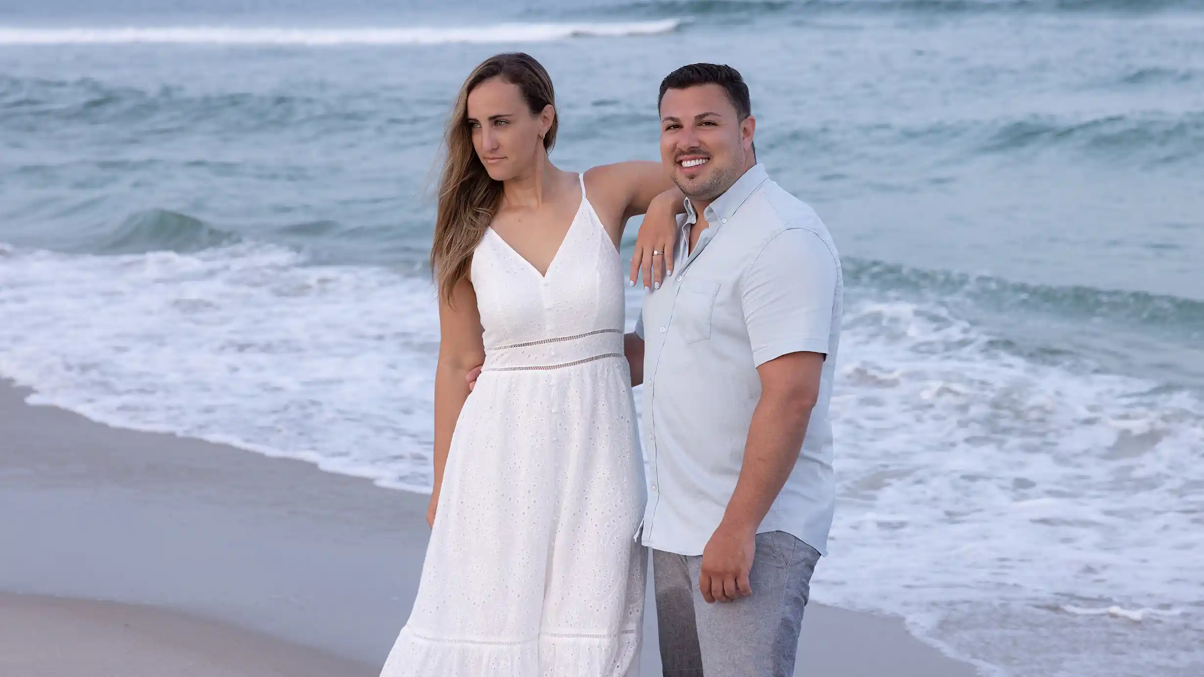 Sunset engagement session at Long Beach Island, NJ. Couple in casual attire standing in the sand next to the ocean.