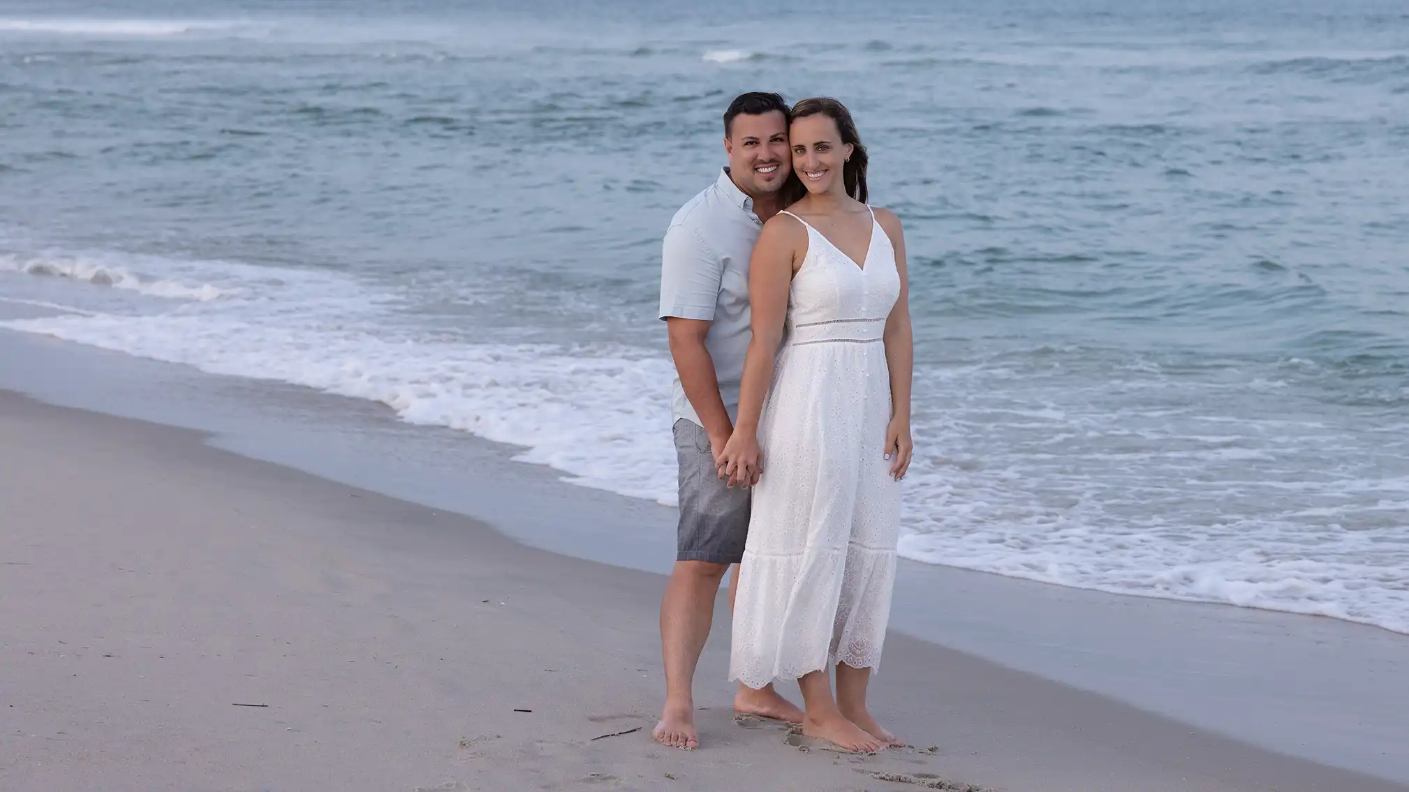 Sunset engagement session at Long Beach Island, NJ. Couple in casual attire standing in the sand next to the ocean.