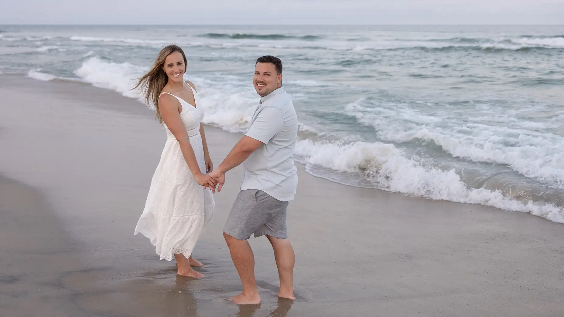 Sunset engagement session at Long Beach Island, NJ. Couple in casual attire standing in the sand next to the ocean.