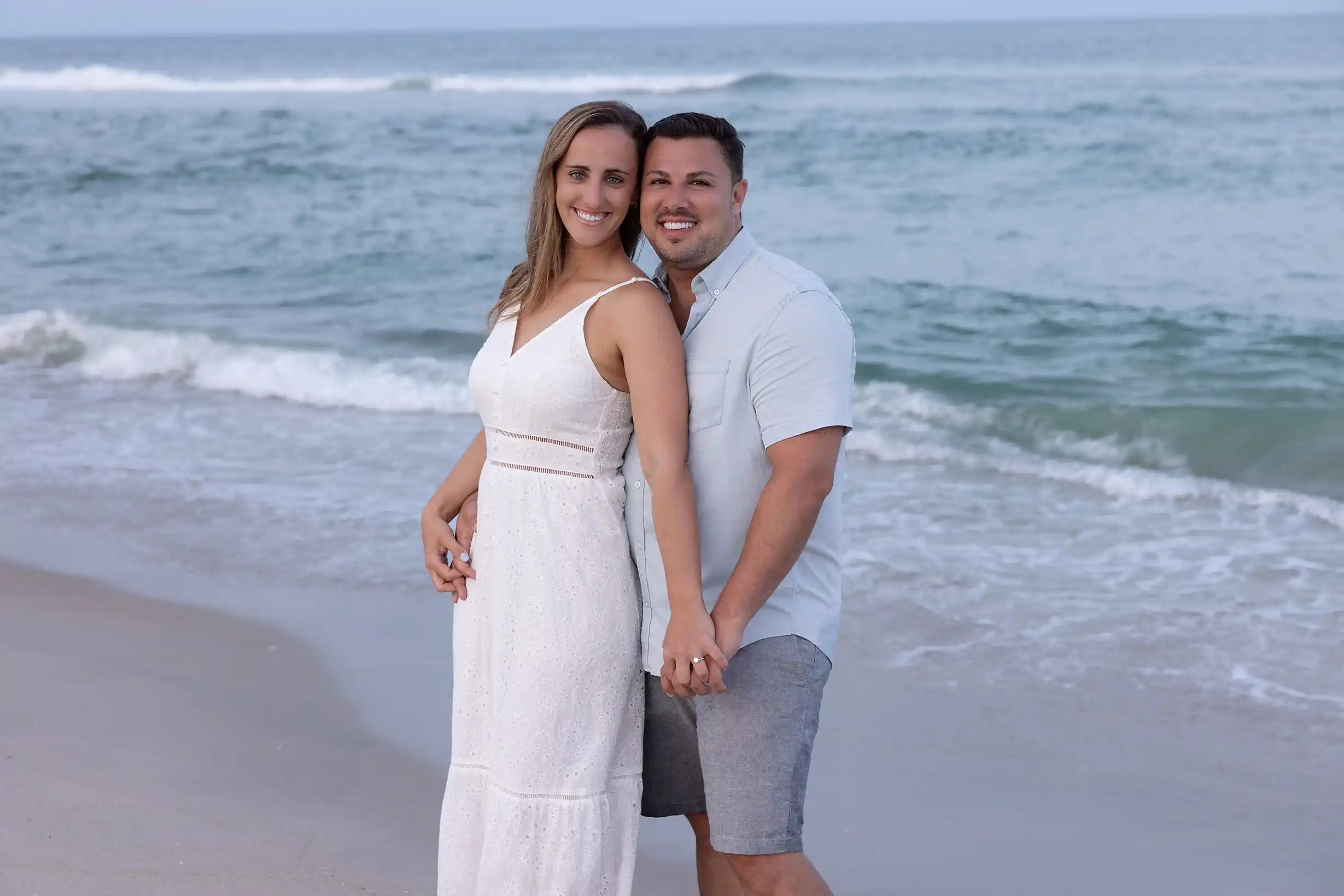 Sunset engagement session at Long Beach Island, NJ. Couple in casual attire standing in the sand next to the ocean.