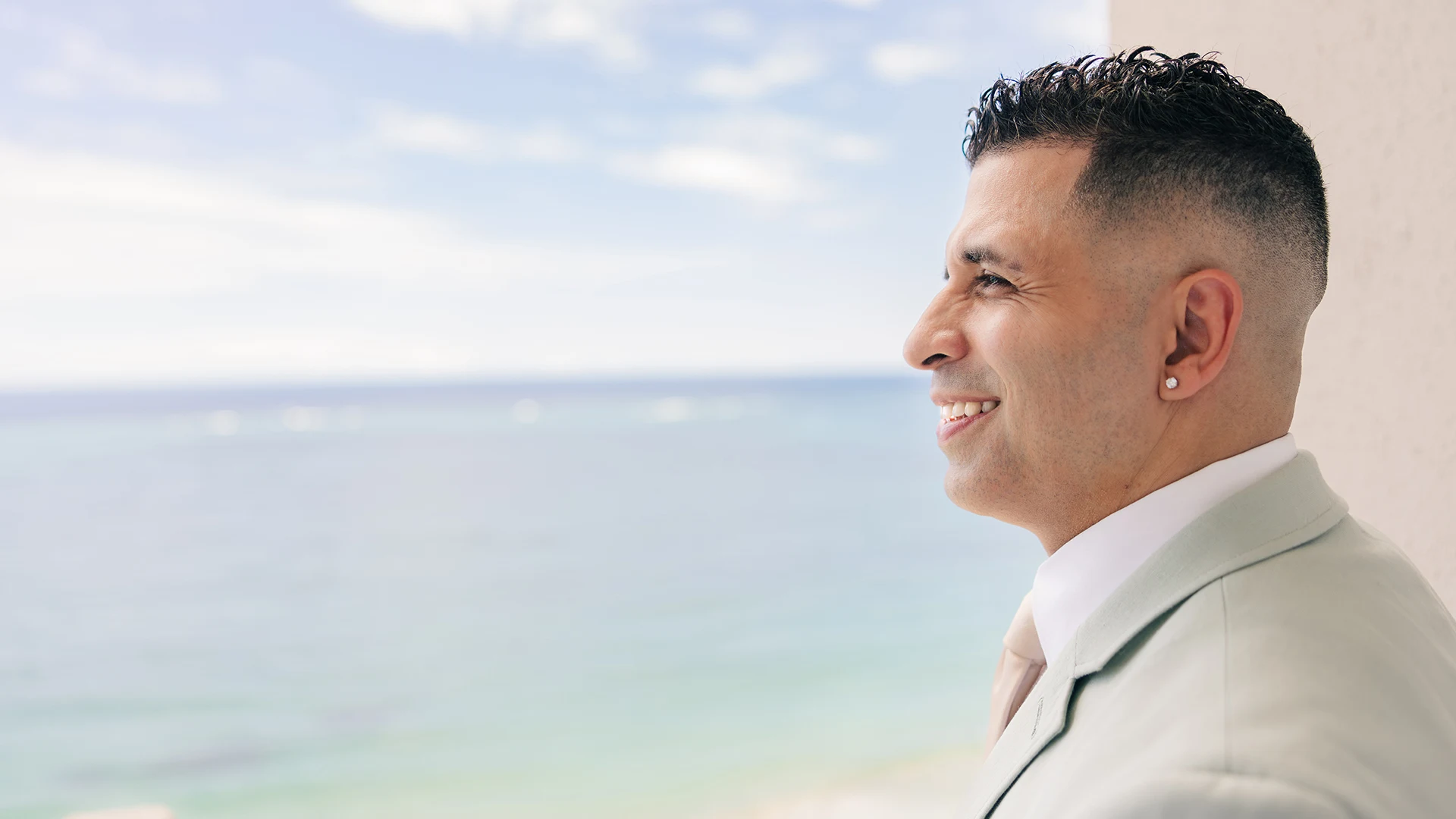 A groom standing outside in Puerto Rico dressed in a suite smiling and looking out at the ocean view exited for his wedding.