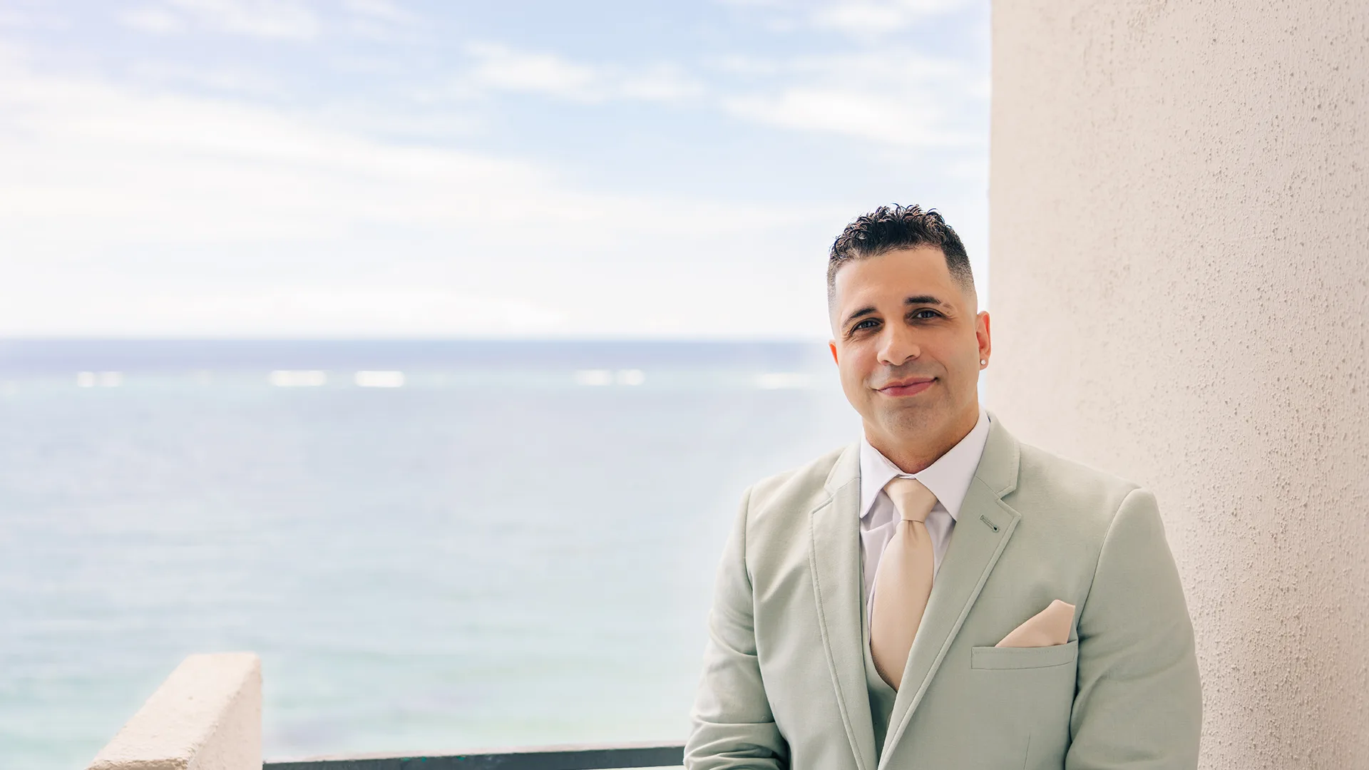 A groom standing outside in Puerto Rico dressed in a suite with an ocean-view background exited for his wedding.