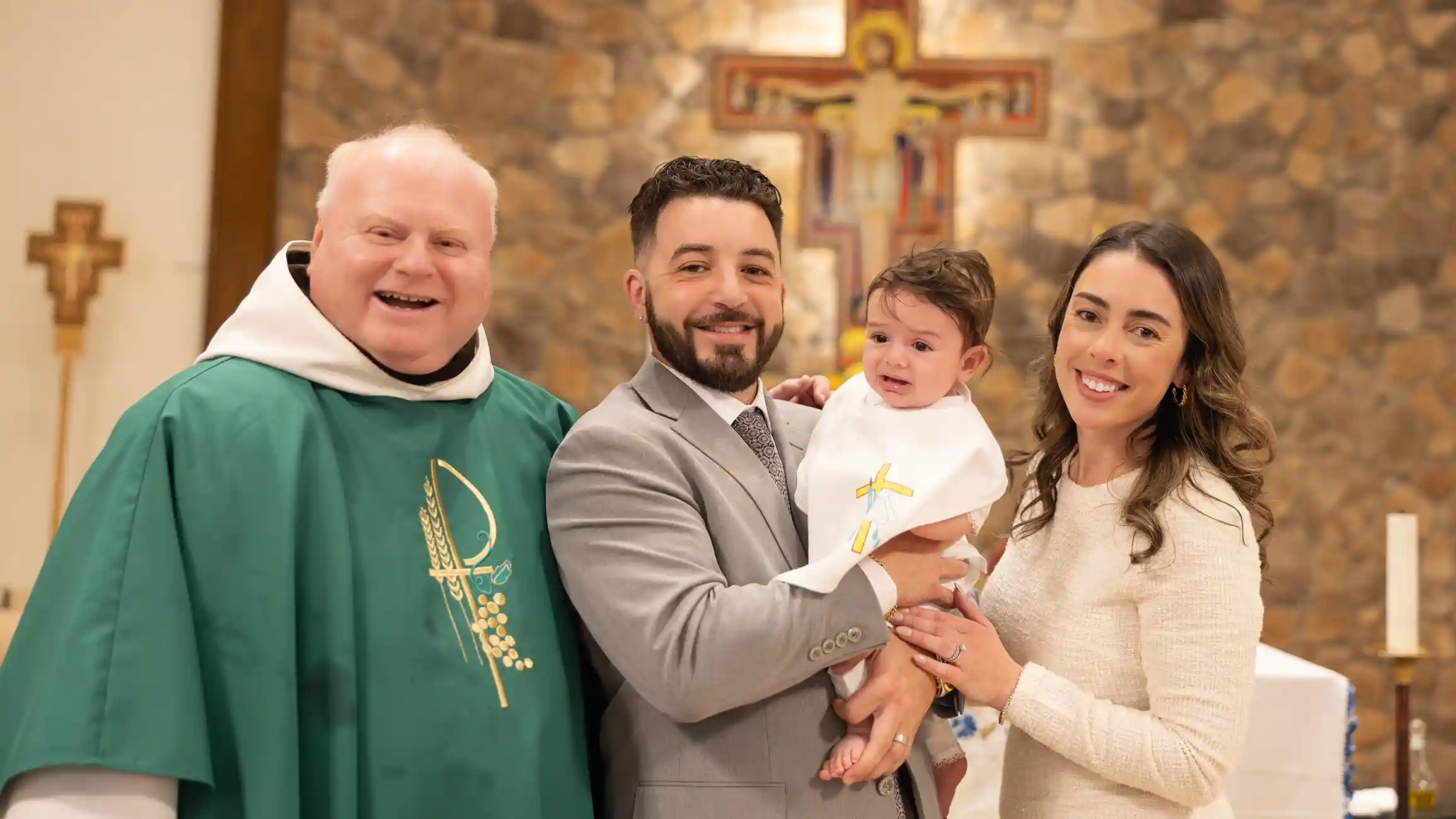 A joyous mother and father holding their son at the alter smiling at the camera with the priest.