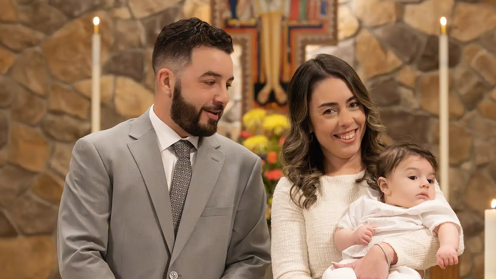 A joyous mother and father holding their son at the alter smiling at the camera.