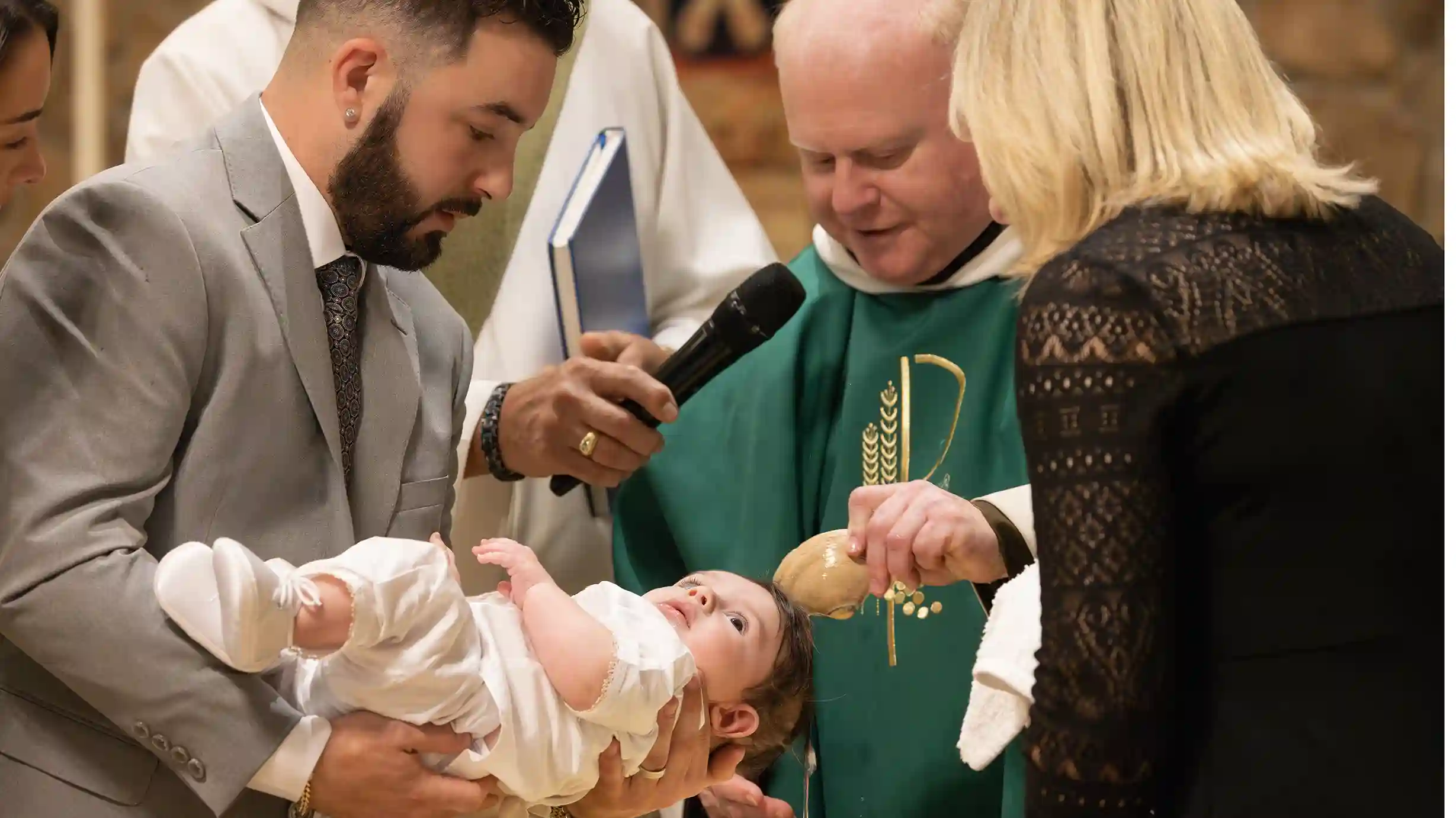 A baby boy being baptized by a priest being held by his father and mother smiling.