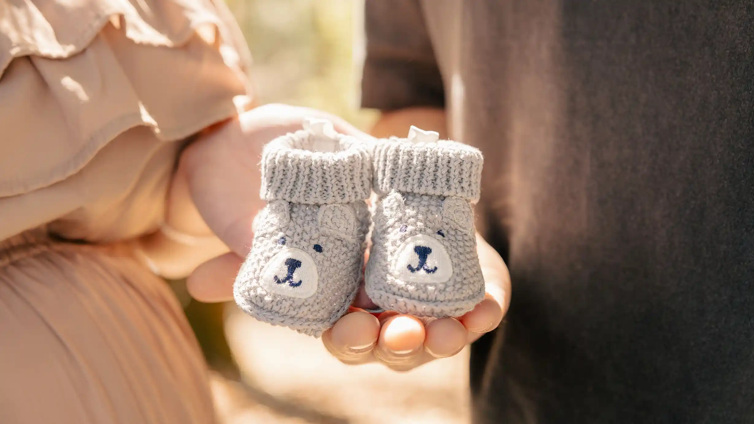 Excited parents-to-be showcasing their baby's shoes in a beautiful natural setting on a bright day.