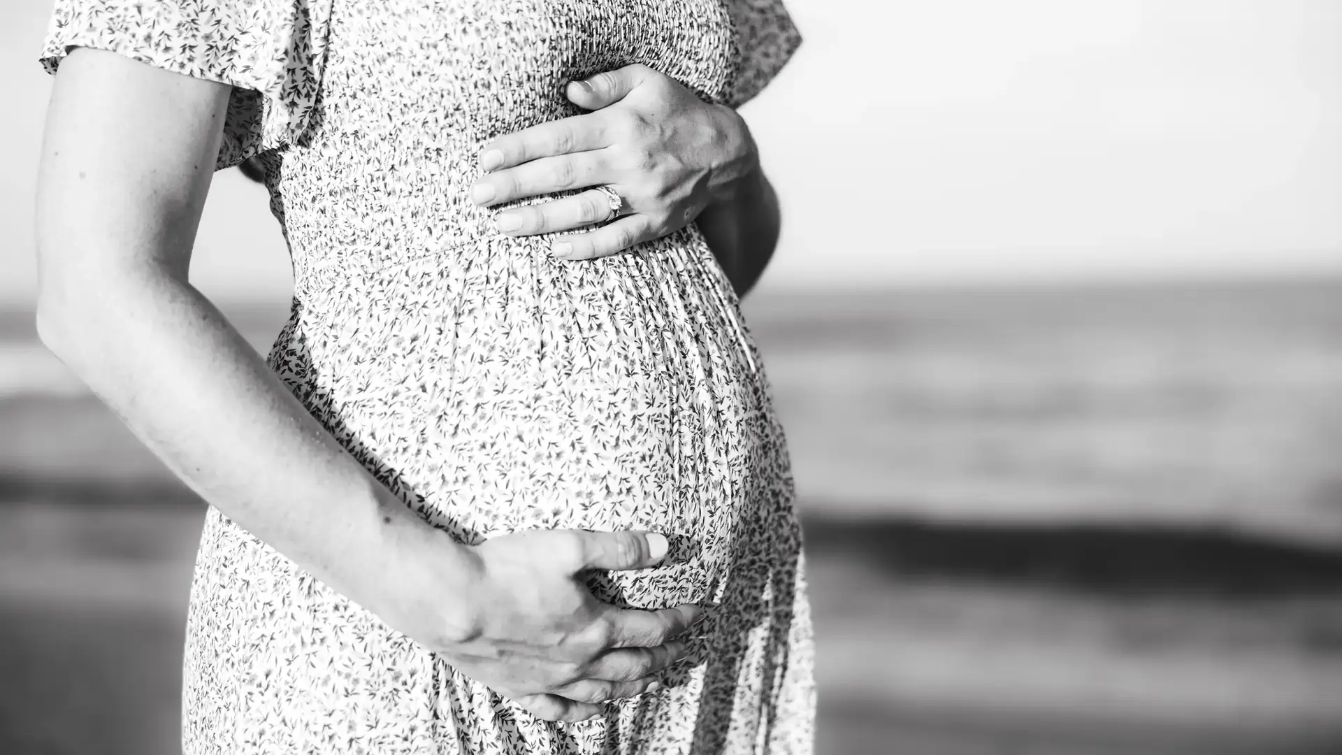 Black and white photo of a pregnant woman in a flowing sun dress, gently cradling her belly on a beach. The vast ocean stretches out behind her.