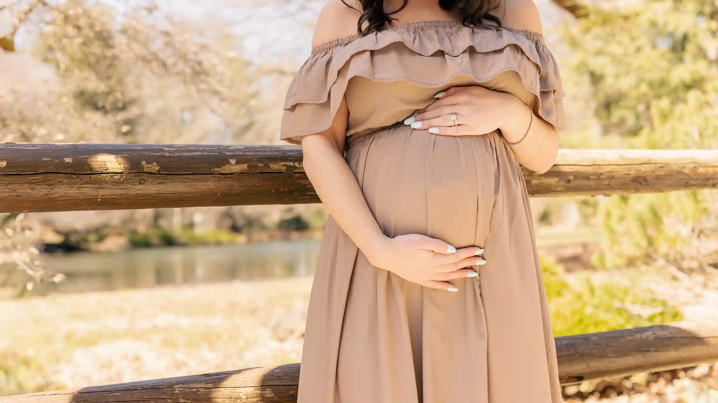 A wife, glowing with anticipation, cradles her baby bump amidst the beauty of nature on a bridge.