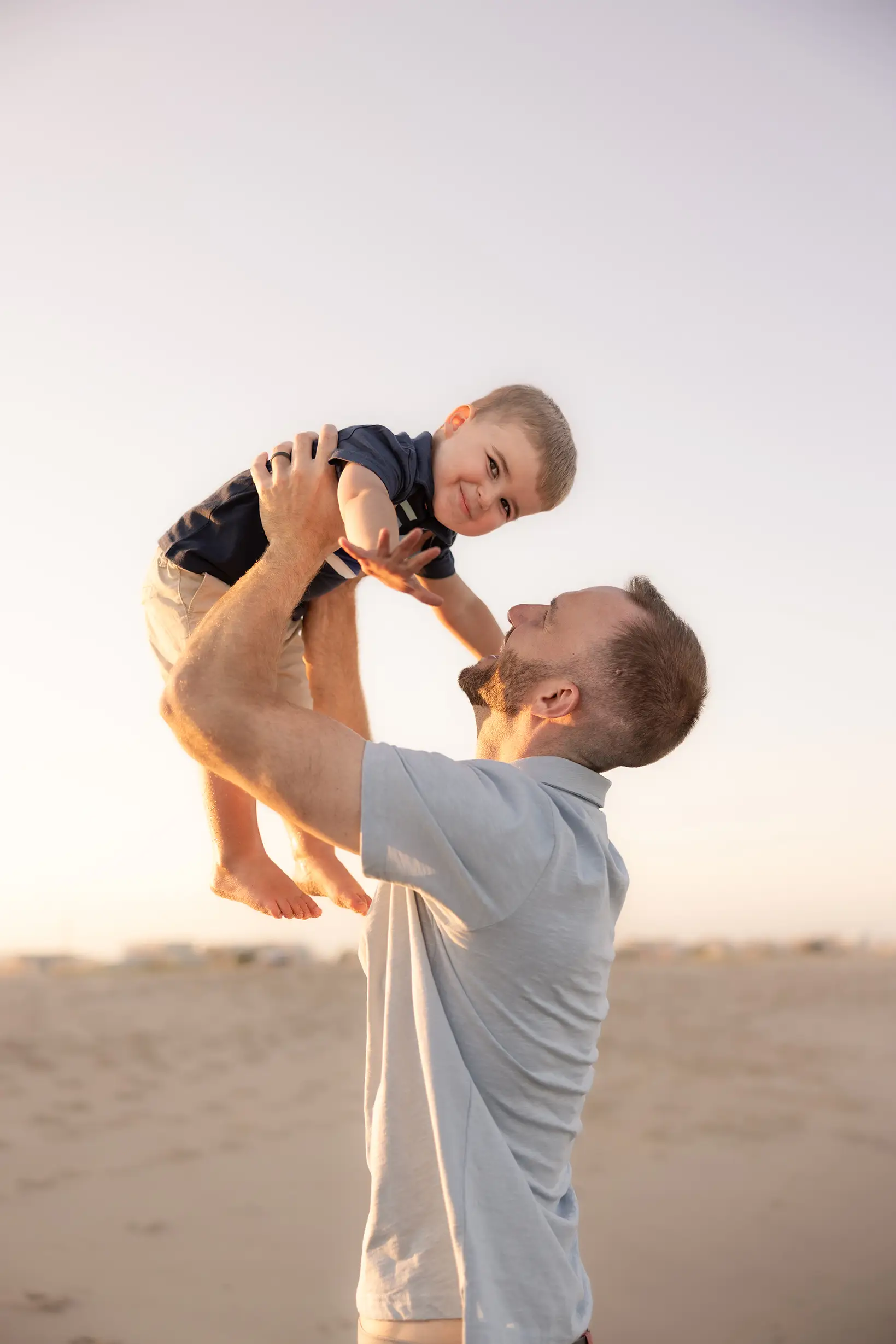 A little boy with his father being tossed and pretending to fly like an airplane with a smile looking at the camera.