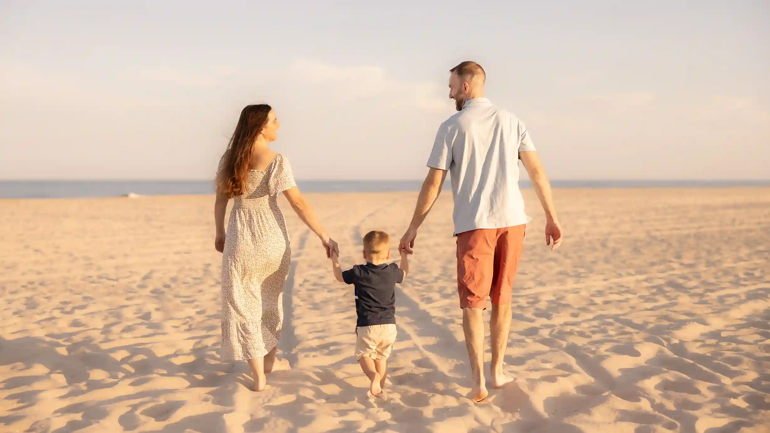 Excited parents-to-be holding the hands of their little boy while walking at the beach.