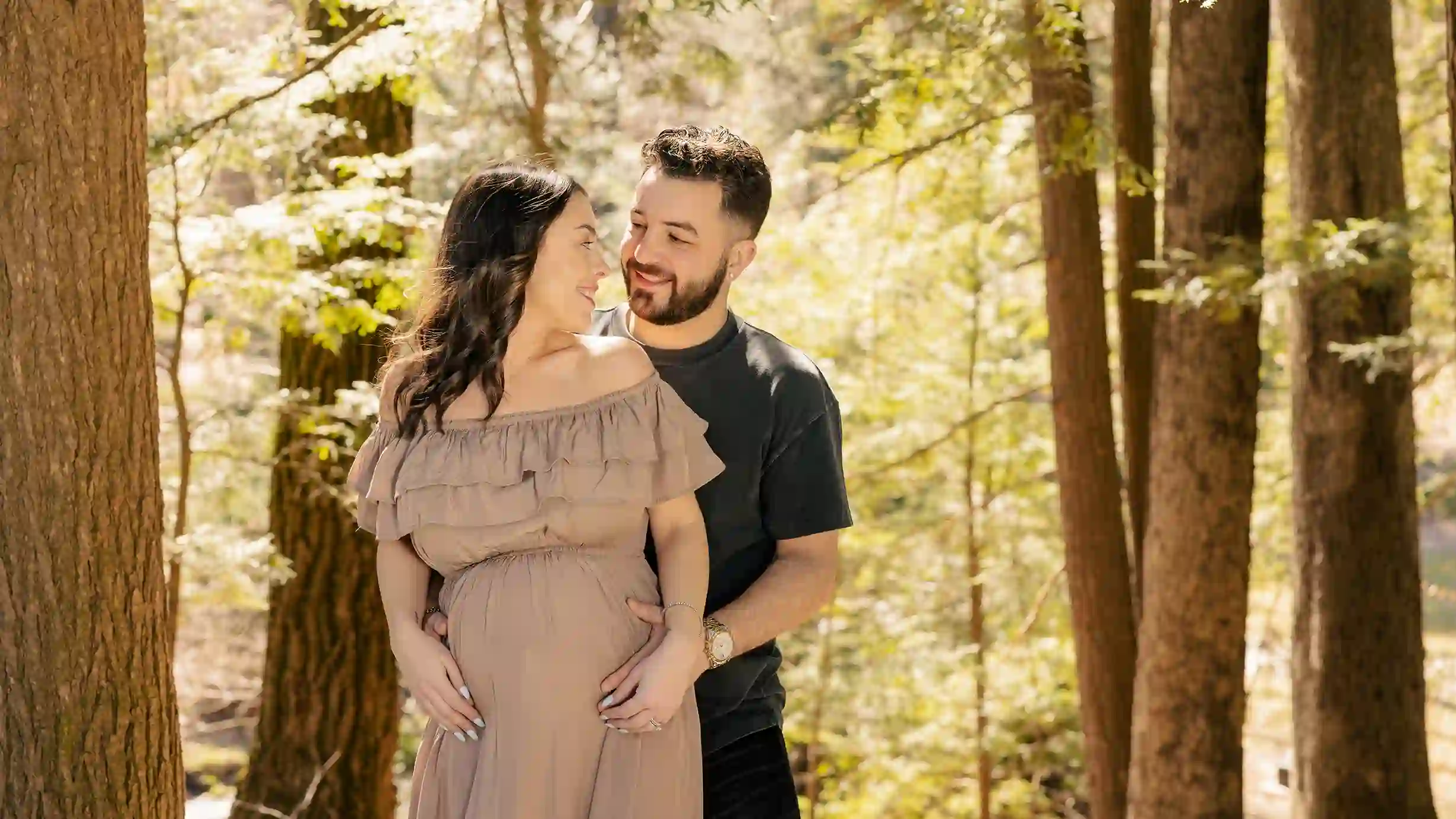 A glowing expectant mother and father, filled with excitement, share a loving look amidst a beautiful wooded area.