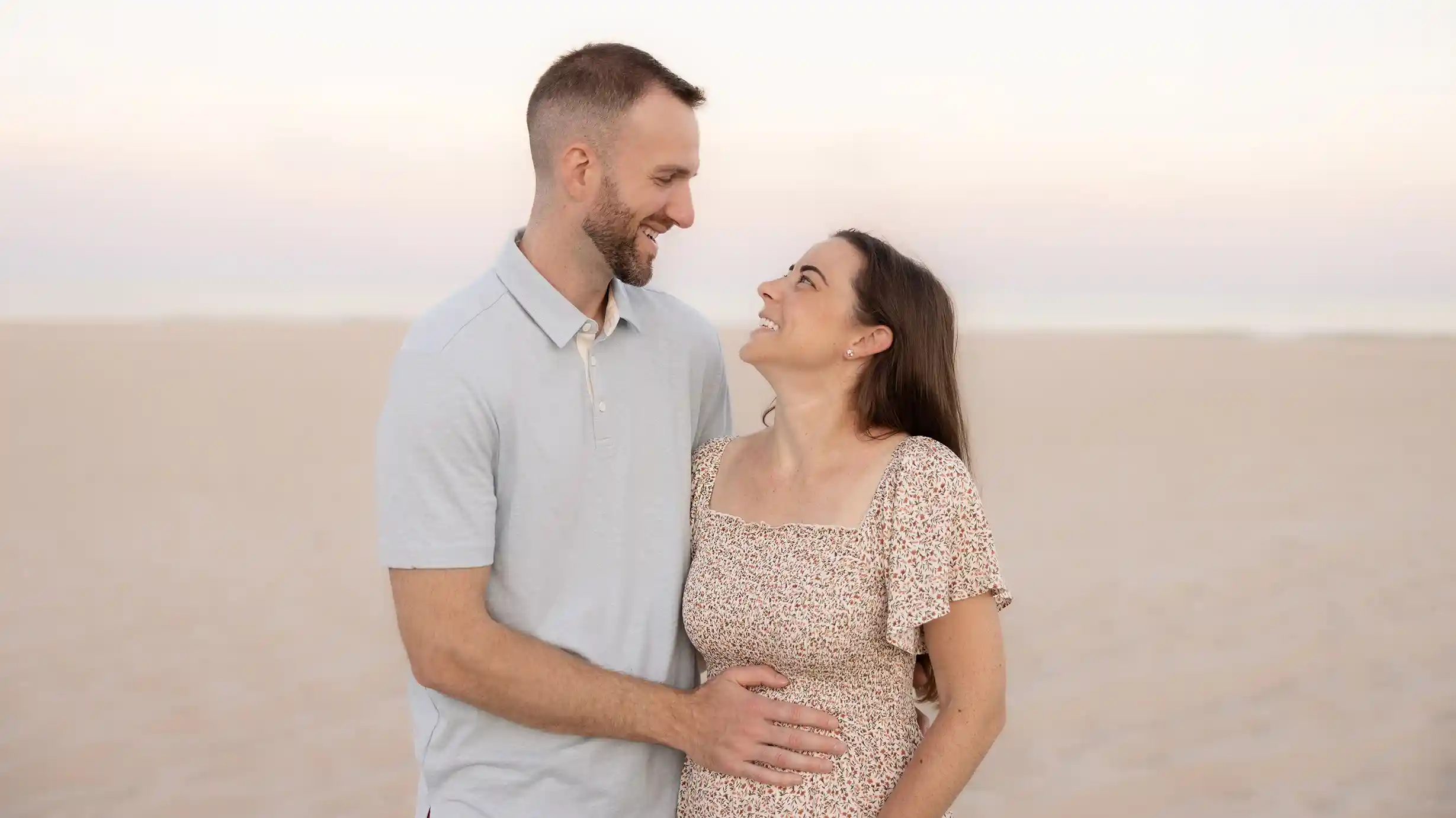 Excited parents-to-be, husband filled with joy as he touches his pregnant wife's belly with the beautiful beach behind them.