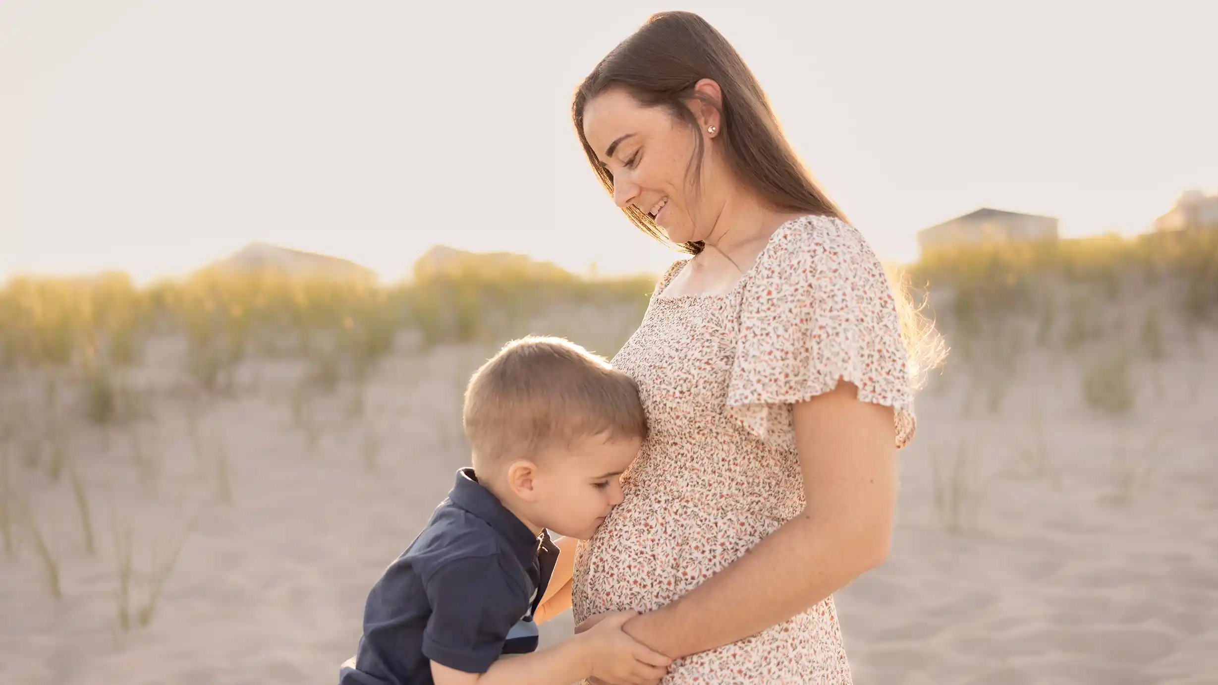 A little boy kissing his mothers belly on a sunny New Jersey beach.