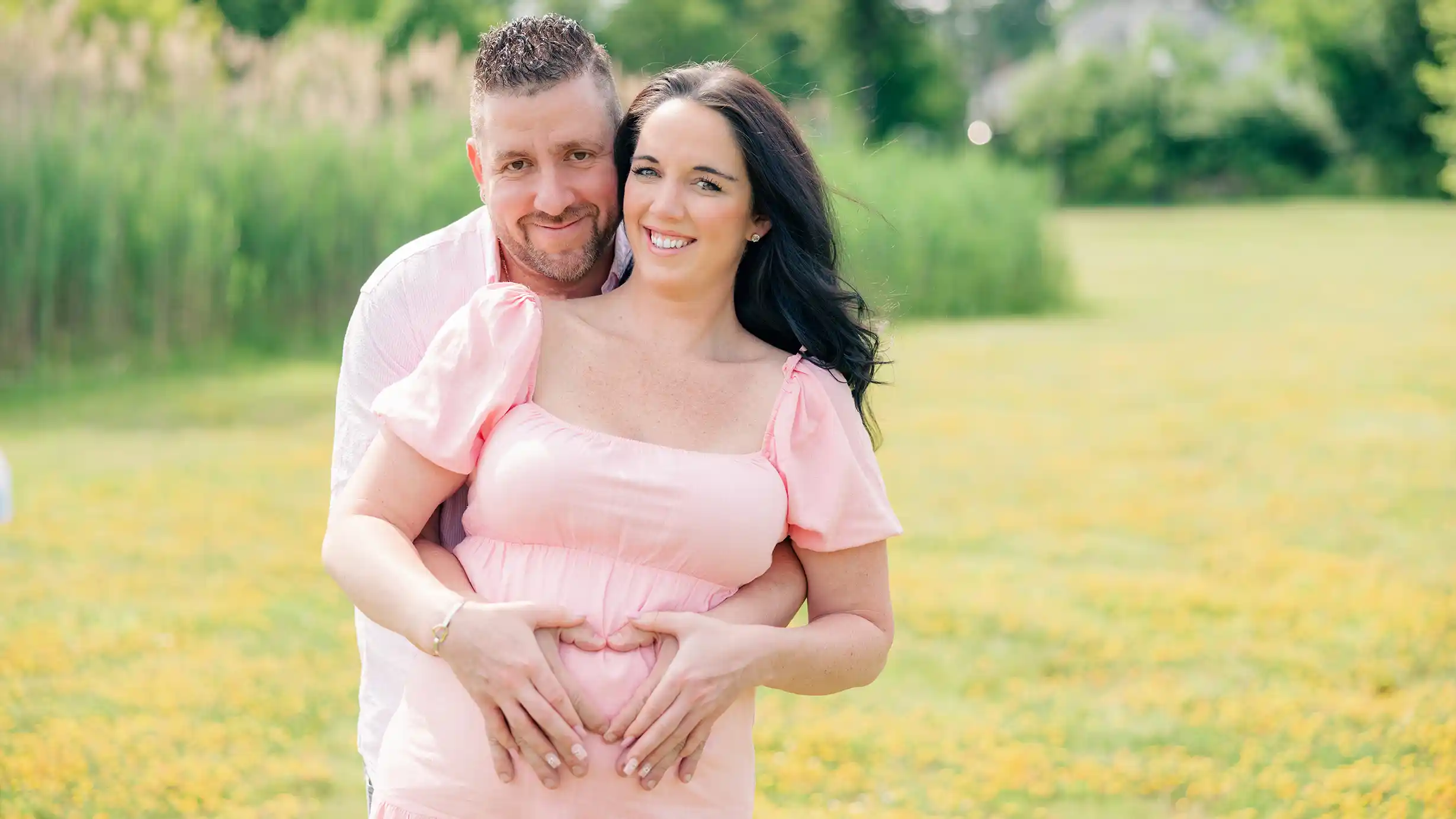 Happy couple expecting a baby, create a heart symbol over the mother's belly in a scenic field.