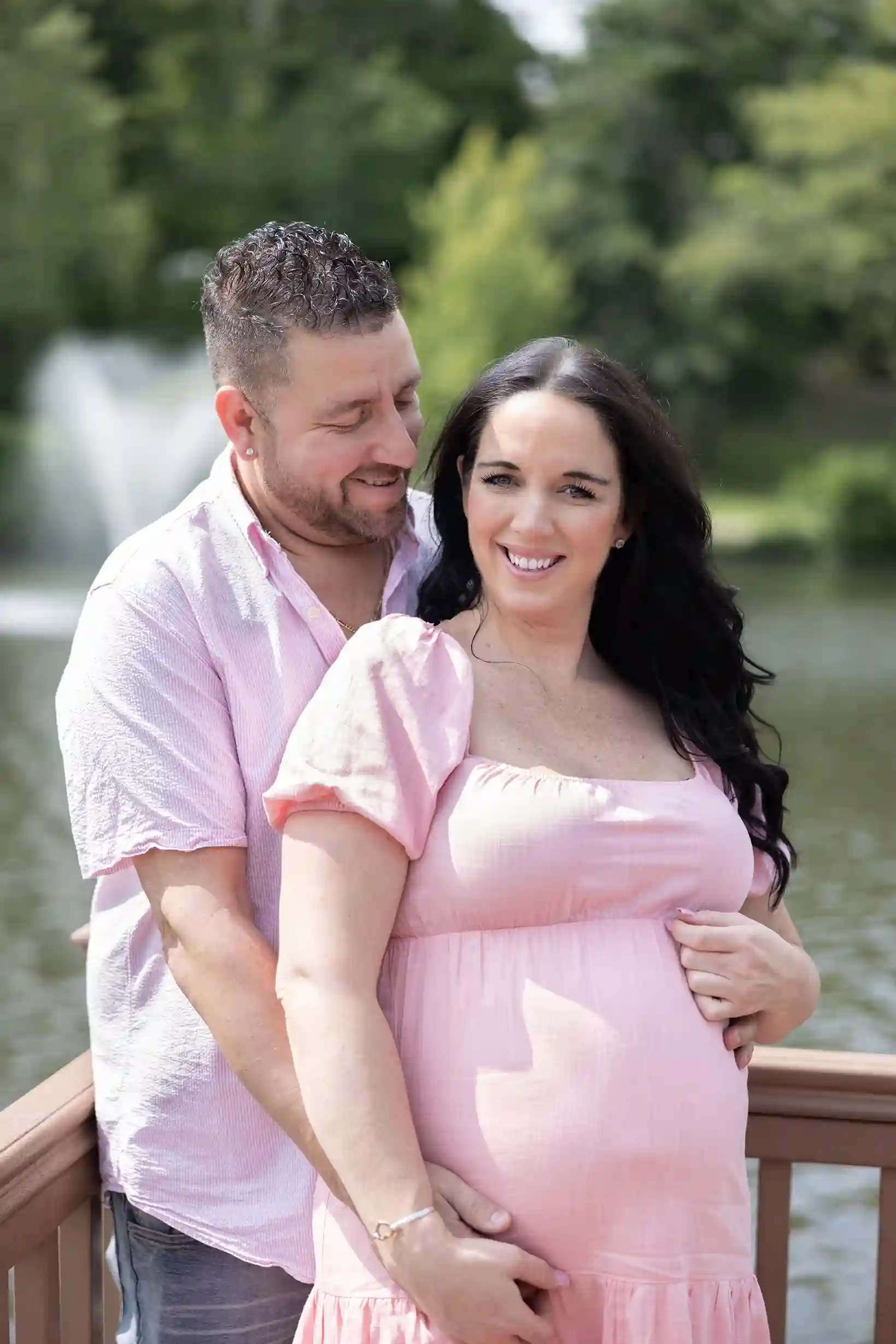 Wife embraced by her husband with a picturesque pond and fountain in the background.