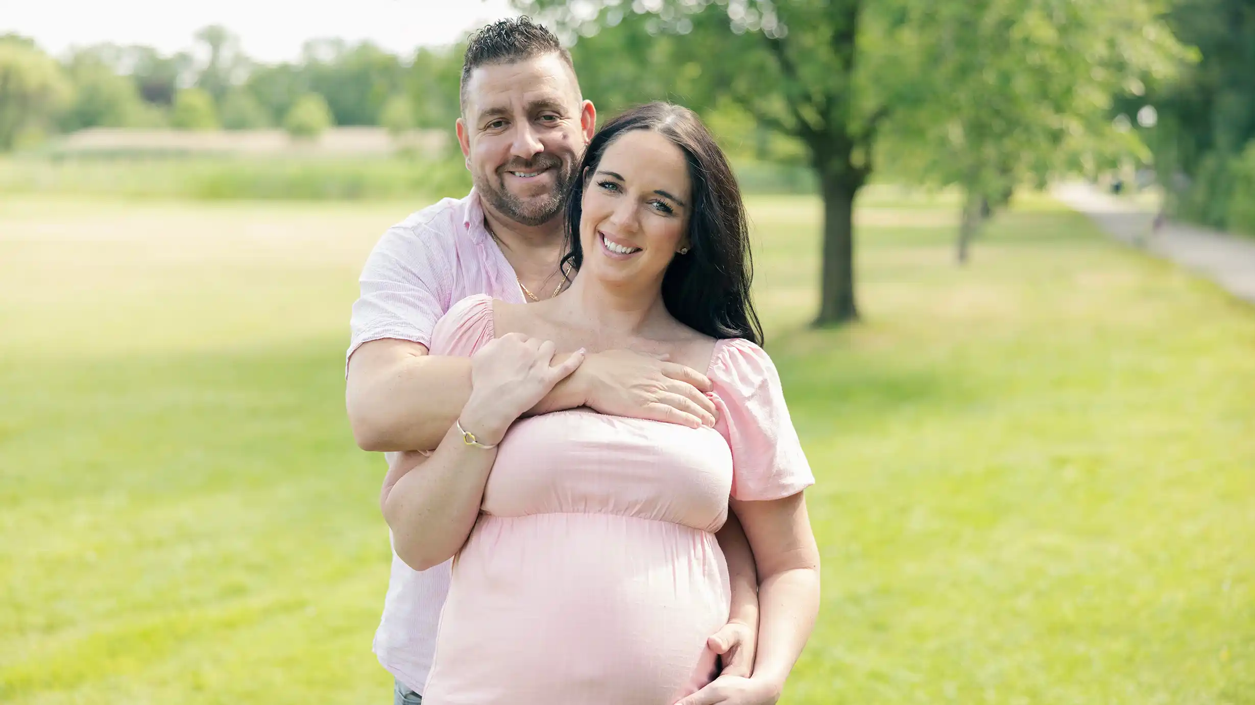 A husband wraps his arms around his wife, holding her close in a lush green field.