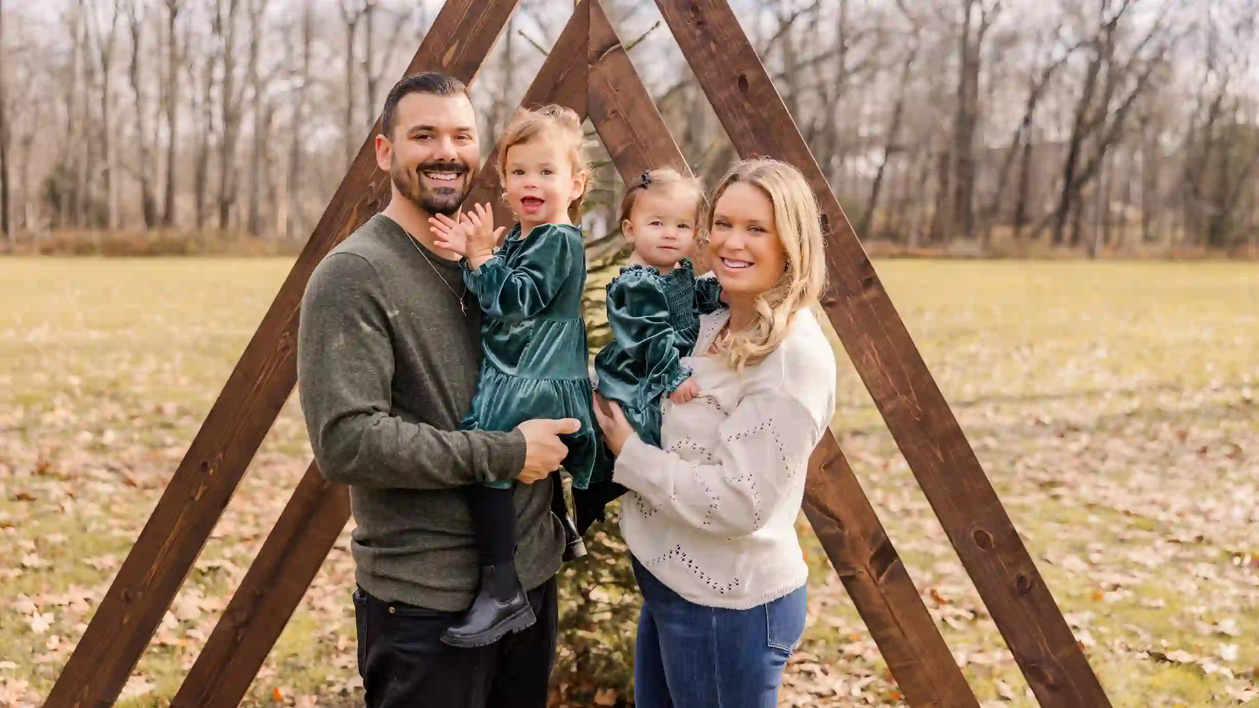 A beautiful family at a local park with a wooden Christmas tree structure with a mini Christmas tree happy, clapping and smiling.