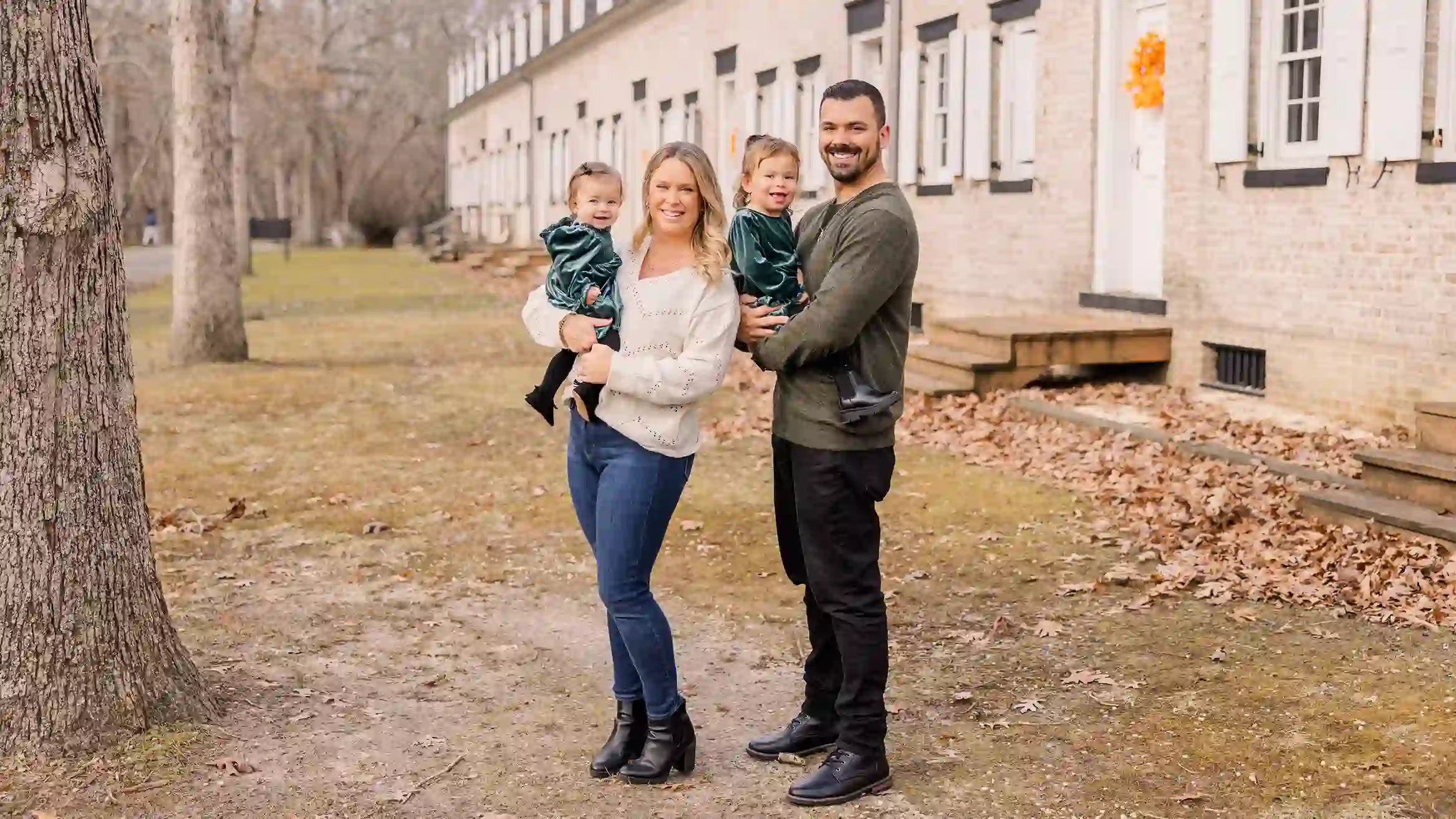 A family with two little girls posing for their Christmas photoshoot in a local park smiling and happy with beautiful fall foliage in the background.