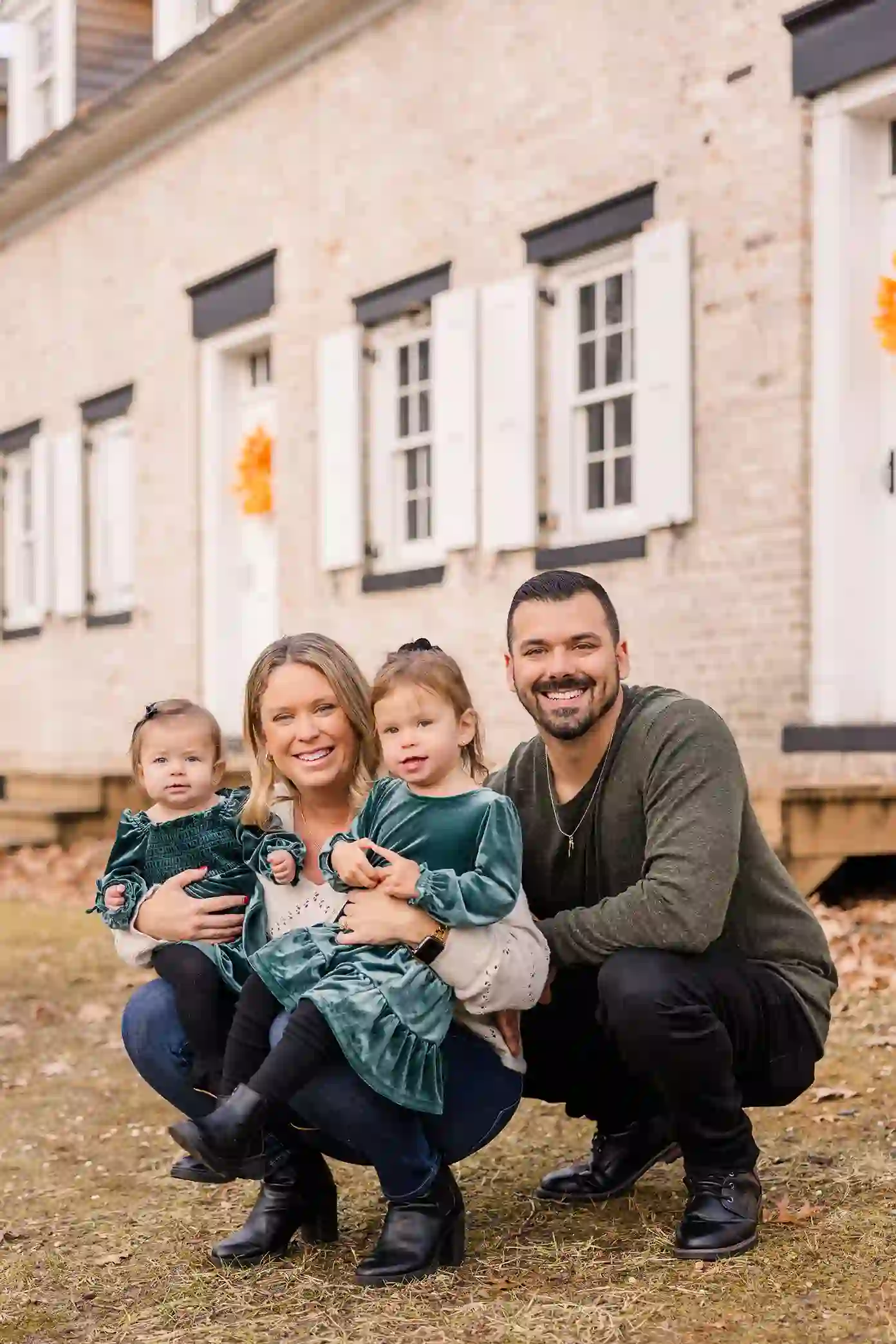 Two beautiful little girls with mom and dad smiling infant of a decorated building with fall foliage.