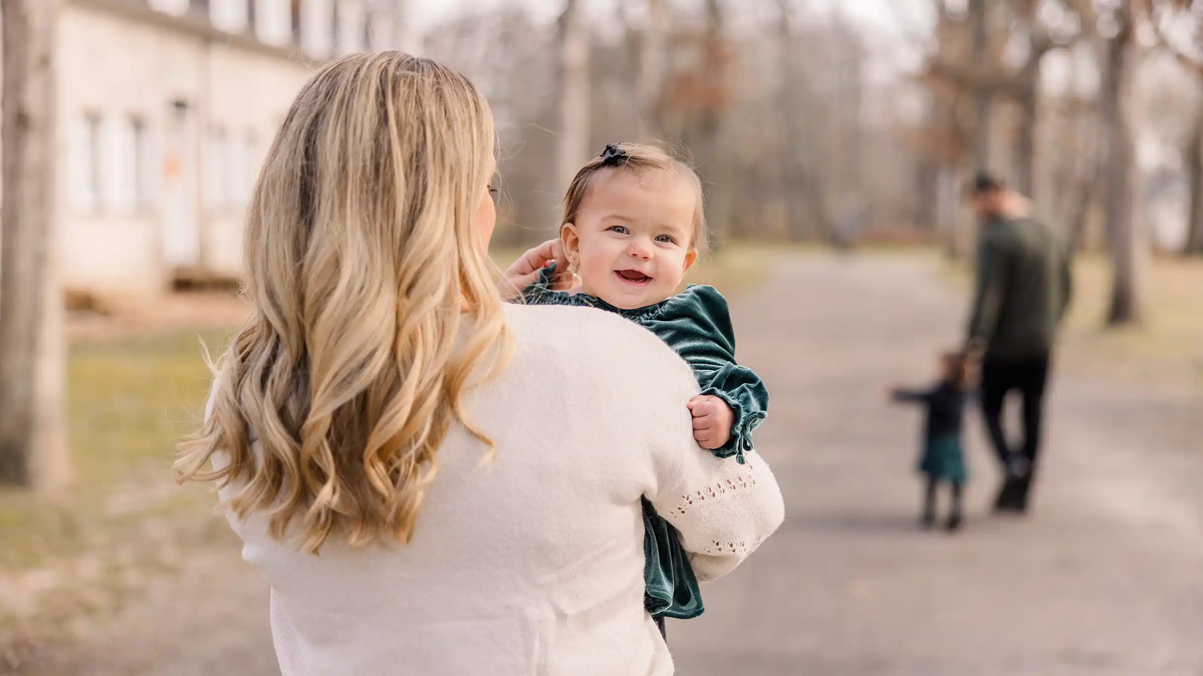 A little baby girl being carried by her mom walking through the park while happy and snuggling with each other while dad walks with the sister in the distance.