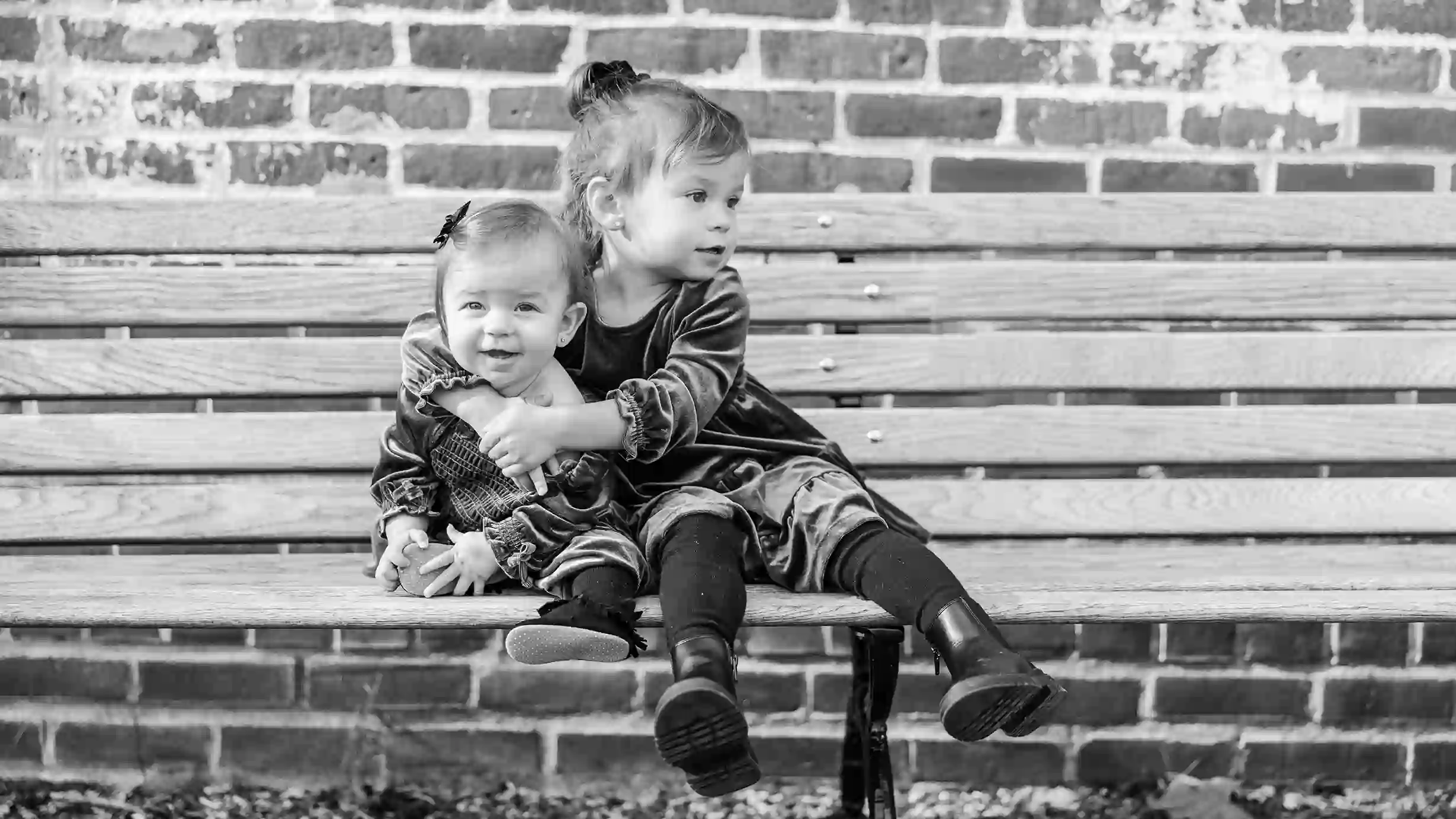 A little girl with her arm over her baby sister while sitting on a park bench while the baby is looking at the camera smiling.