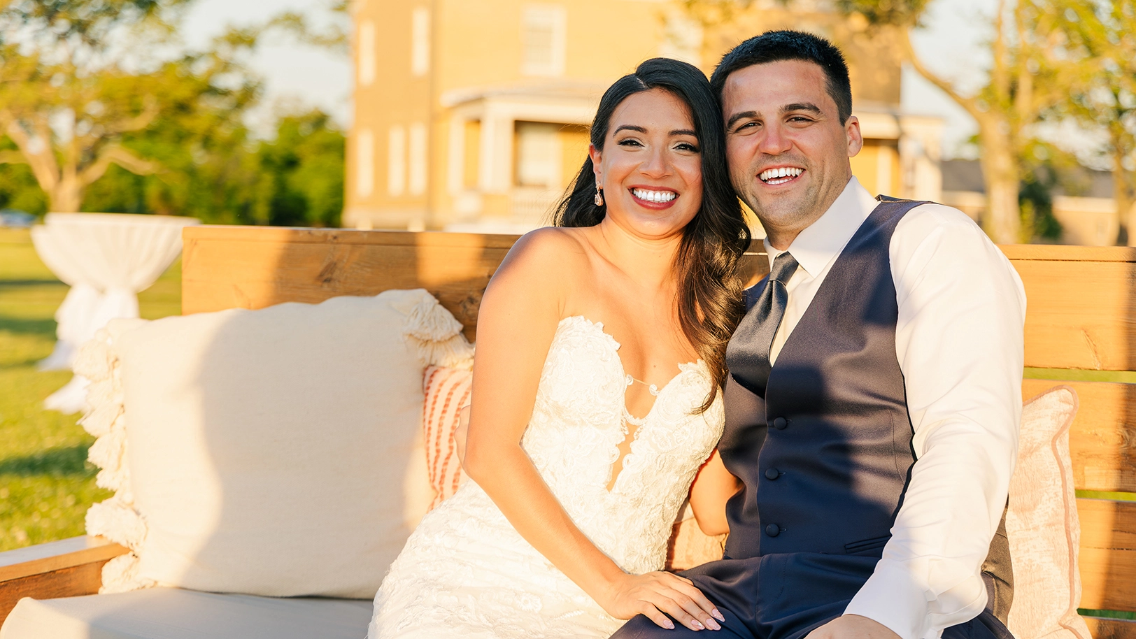 Bride and groom smile for the camera at their outdoor wedding reception with a colorful sunset.