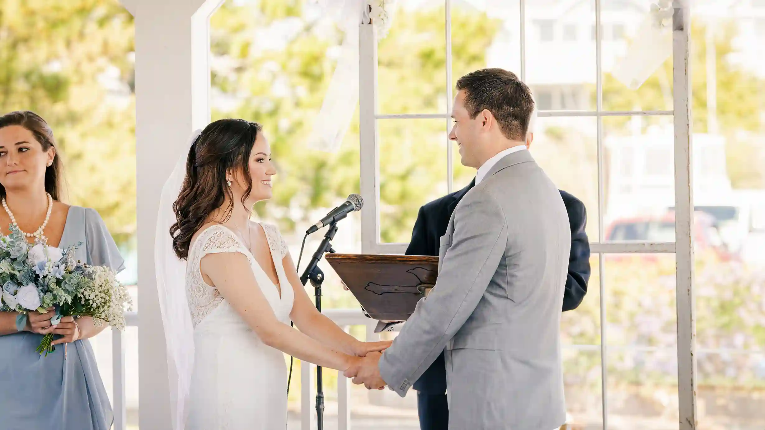 A radiant bride and groom, filled with love, share a smile at the altar while listening to the officiant with the maid of honor watching.