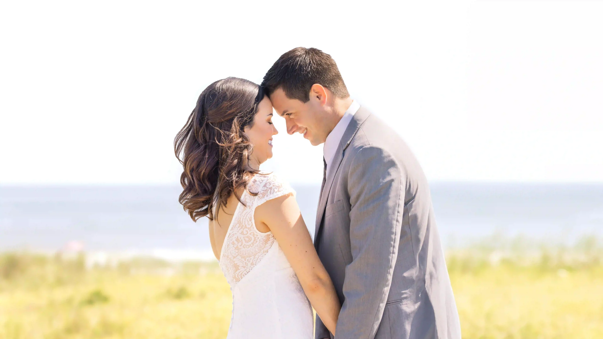 Newlyweds in Barnegat Light, NJ, celebrating on the beach after their wedding ceremony. Bride and groom stand face-to-face at the water's edge.